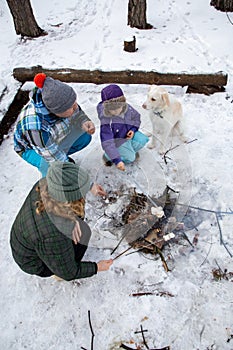 family winter picnic. Happy parents, child, white dog sit around a campfire in the forest and roast marshmallows