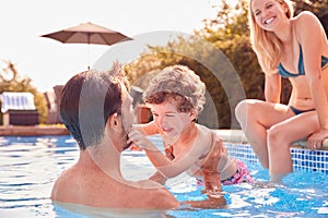 Family With Young Son Having Fun On Summer Vacation Splashing In Outdoor Swimming Pool