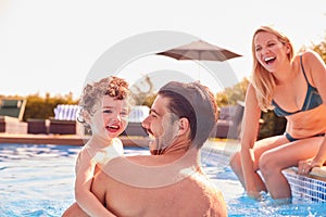 Family With Young Son Having Fun On Summer Vacation Splashing In Outdoor Swimming Pool