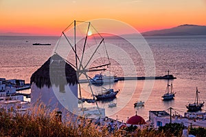 Famous traditional Greek windmill over port of Mykonos, Greece at sunset. Beautiful sky, sun touch sea horizon, boats