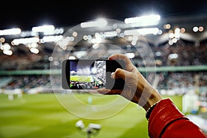 Fan hand with smartphone photographing football game. Using mobile phone camera at the stadium.