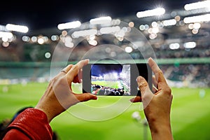 Fan hand with smartphone photographing football game. Using mobile phone camera at the stadium.