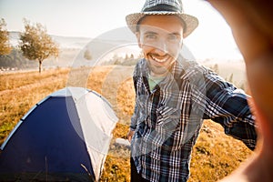 Fanny bearded man smiling and taking selfie in mountains from his smart phone. Traveler man with beard wearing hat take