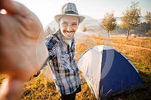 Fanny bearded man smiling and taking selfie in mountains from his smart phone. Traveler man with beard wearing hat take self