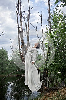 Fantastic fairy beautiful woman in white long dress on old dry tree. Girl princess in swamp. Fashion model sexy girl posing on a