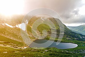 Fantastic wide peaceful view of blue lake in green valley and small tourist tents at rocky mountain with patches of snow covered