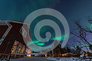 Fantastic winter landscape with wooden house with light in window in snowy mountains and Northern light in night sky. Christmas