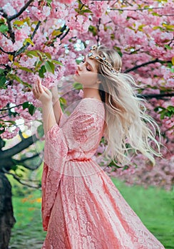 Fantasy happy woman princess walks in spring blooming garden hugs herself by shoulders. Pink flowers sakura tree summer