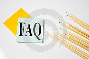 FAQ word written on a yellow piece of paper and white background with pencils lying next to it. text