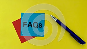 FAQs text on blue post it note with pen and yellow background. Business concept.