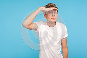 Far away vision. Portrait of inquisitive man in casual white t-shirt peering long distance and holding hand over his eyes