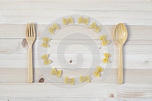 Farfalle arranged in a circle with spoon and fork on light pine wood background.