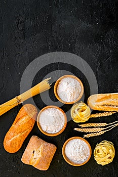 Farinaceous food. Fresh bread and raw pasta near flour in bowl and wheat ears on black background top view copy space