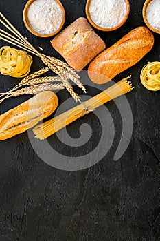 Farinaceous food. Fresh bread and raw pasta near flour in bowl and wheat ears on black background top view copy space photo