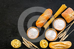Farinaceous food. Fresh bread and raw pasta near flour in bowl and wheat ears on black background top view copy space