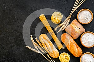 Farinaceous food. Fresh bread and raw pasta near flour in bowl and wheat ears on black background top view copy space