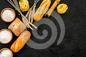 Farinaceous food. Fresh bread and raw pasta near flour in bowl and wheat ears on black background top view copy space