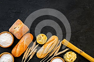 Farinaceous food. Fresh bread and raw pasta near flour in bowl and wheat ears on black background top view copy space