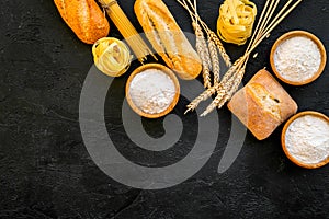 Farinaceous food. Fresh bread and raw pasta near flour in bowl and wheat ears on black background top view copy space