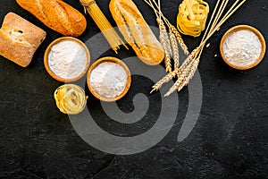 Farinaceous food. Fresh bread and raw pasta near flour in bowl and wheat ears on black background top view copy space