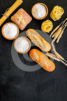 Farinaceous food. Fresh bread and raw pasta near flour in bowl and wheat ears on black background top view space for photo