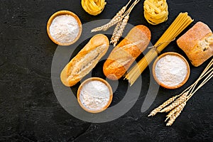 Farinaceous food. Fresh bread and raw pasta near flour in bowl and wheat ears on black background top view space for