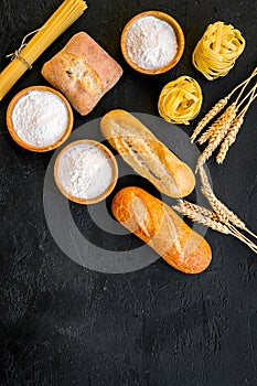 Farinaceous food. Fresh bread and raw pasta near flour in bowl and wheat ears on black background top view space for