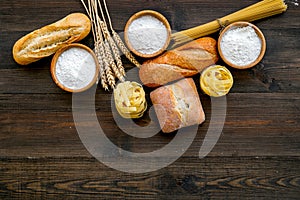 Farinaceous food. Fresh bread and raw pasta near flour in bowl and wheat ears on dark wooden background top view space