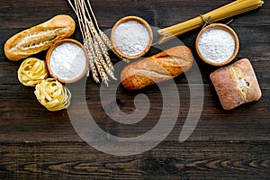 Farinaceous food. Fresh bread and raw pasta near flour in bowl and wheat ears on dark wooden background top view space