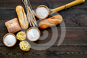 Farinaceous food. Fresh bread and raw pasta near flour in bowl and wheat ears on dark wooden background top view space