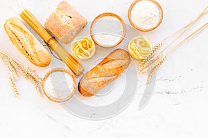 Farinaceous food. Fresh bread and raw pasta near flour in bowl and wheat ears on white stone background top view copy