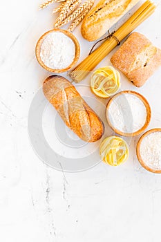 Farinaceous food. Fresh bread and raw pasta near flour in bowl and wheat ears on white stone background top view copy