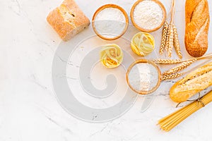 Farinaceous food. Fresh bread and raw pasta near flour in bowl and wheat ears on white stone background top view space