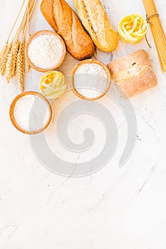 Farinaceous food. Fresh bread and raw pasta near flour in bowl and wheat ears on white stone background top view space