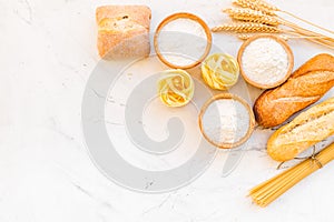 Farinaceous food. Fresh bread and raw pasta near flour in bowl and wheat ears on white stone background top view space