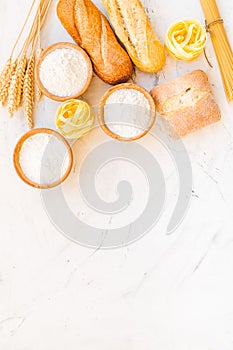 Farinaceous food. Fresh bread and raw pasta near flour in bowl and wheat ears on white stone background top view space photo