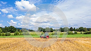 A farm tractor cultivating a harvested field against  a blue sky with clouds on a sunny summer day