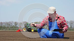 Farmer, agronomist sits between soil rows, checking quality of soil on farm field. background of working tractor