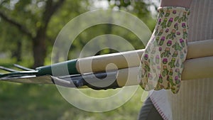 The farmer in garden gloves walking through the garden with a shovel and pitchfork in hands close-up. Concept of rural