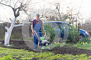 A farmer in the garden tills the land with a motorized cultivator or power tiller, preparing the soil for planting crops