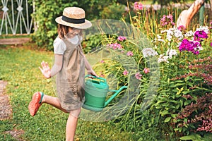 Farmer girl in summer straw hat. Little gardener farming, watering flowerbed with pink flowers, having fun in garden. Big green