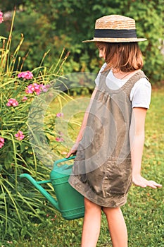 Farmer girl in summer straw hat. Little gardener farming, watering flowerbed with pink flowers, having fun in garden. Big green