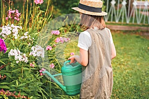 Farmer girl in summer straw hat. Little gardener farming, watering flowerbed with pink flowers, having fun in garden. Big green