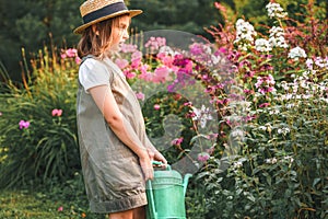 Farmer girl in summer straw hat. Little gardener farming, watering flowerbed with pink flowers, having fun in garden. Big green