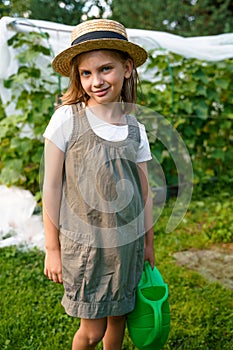 Farmer girl in summer straw hat. Little vegetables gardener farming in garden. Big green watering can water fresh