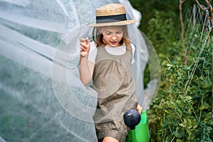 Farmer girl in summer straw hat. Little vegetables gardener farming in garden. Big green watering can water fresh