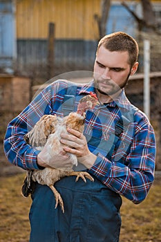 Farmer handsome serious european caucasian rural portrait in countryside with beard, shirt and overalls looks at chicken with