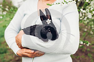 Farmer holding black rabbit in spring garden. Little bunny with flowers on head sitting in hands