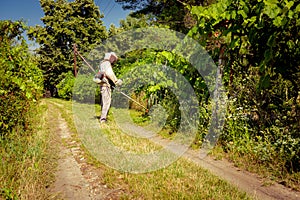 Farmer in protective clothing is mowing grass in garden with manually lawn trimmer