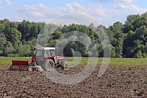 A farmer on a red tractor with a seeder sows grain in plowed land in a private field in the village area. Mechanization of spring